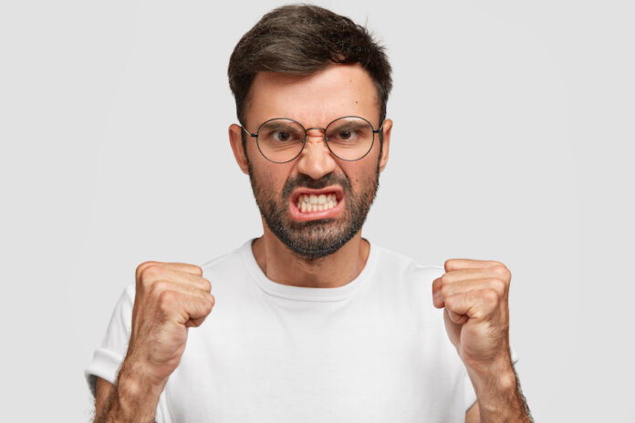 Furious angry European male clenches teeth and fists with rage, tries to control his negative emotions, frowns face in anger, expresses annoyance during quarrel isolated over white background